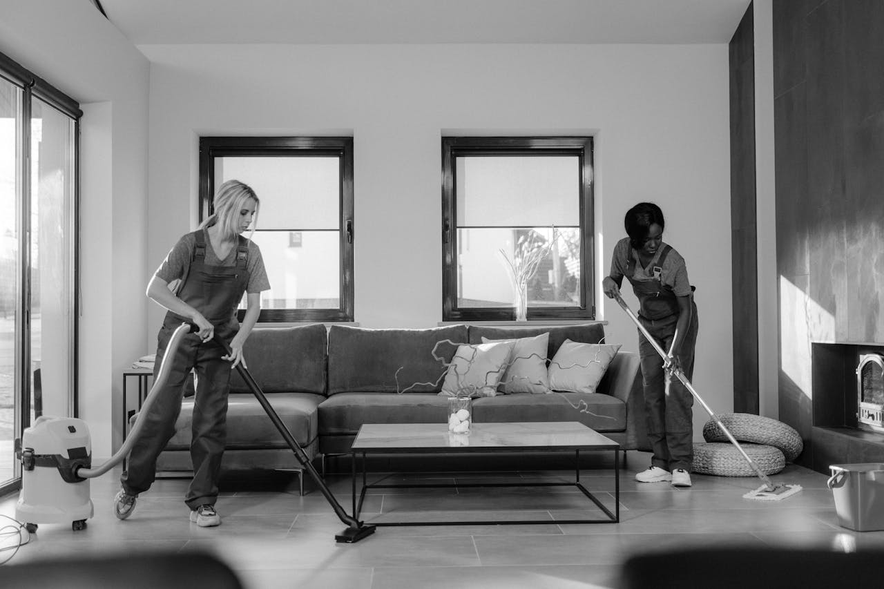 services-04 Two women in work clothes cleaning a modern living room using a vacuum and mop.